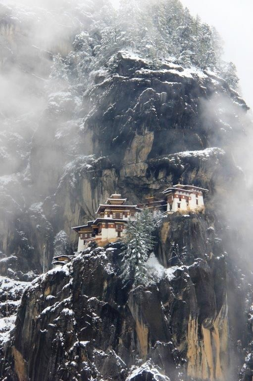 the image depicts the tiger's nest monastery (paro taktsang) perched on a steep, rocky cliff in bhutan during winter. the monastery complex is a cluster of buildings with white walls and brown roofs. snow covers the rocks and trees surrounding the monastery. fog or mist shrouds parts of the mountain, adding to the mystical atmosphere.