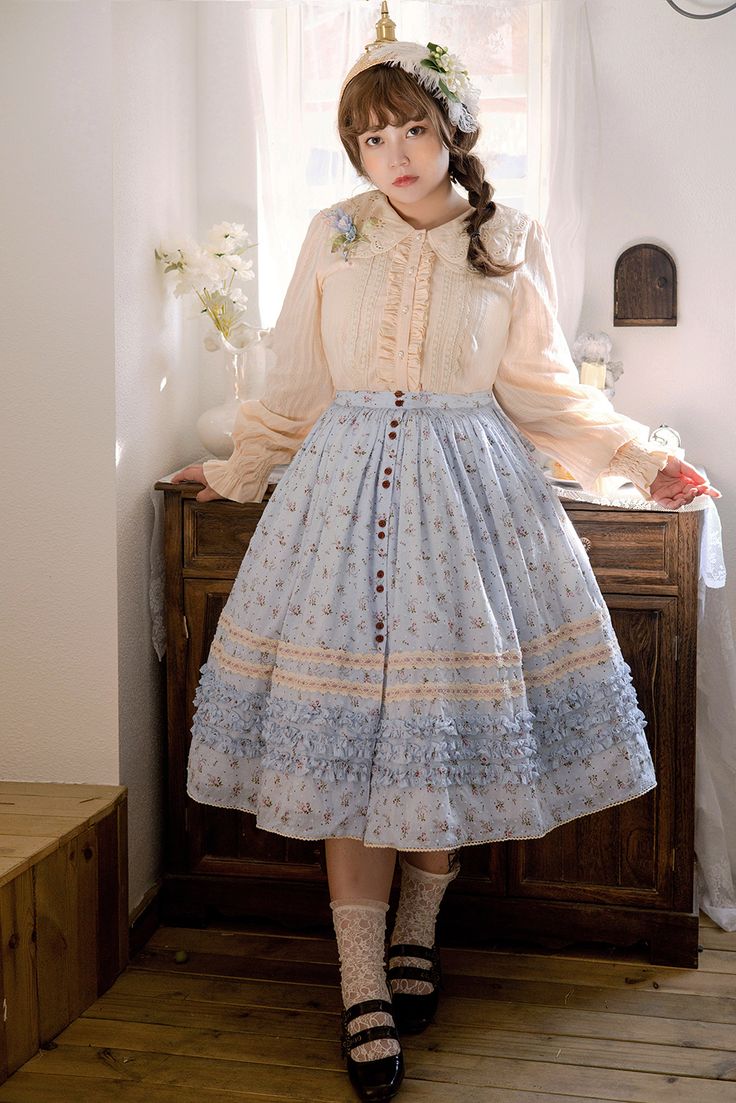 a young woman is posing in a vintageinspired outfit. she is wearing a light blue floral skirt with lace detailing and buttons down the front, paired with a creamcolored, ruffled blouse. her hair is styled in a side braid with a flower crown, and she is wearing white socks and black mary jane shoes. the room she is in has wooden floors and cabinets, with a window letting in bright natural light.