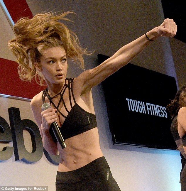 a woman is doing a boxing exercise with her arm extended outward, holding a microphone in her other hand. she is wearing a black sports bra and black pants, and has her blonde hair pulled back in a ponytail. she is in a gym or workout space and is surrounded by a red wall and a tv with the logo "tough fitness" on it.