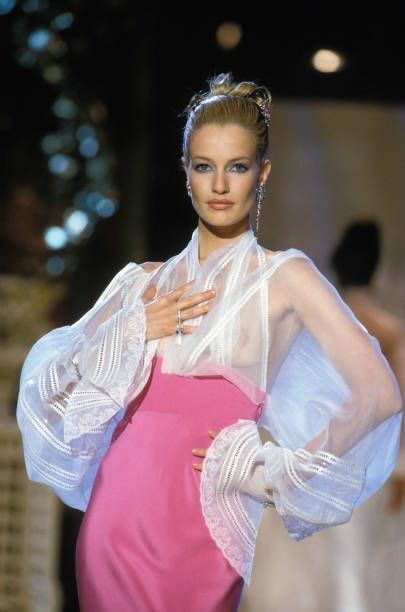 a model wearing a pink dress with a sheer white blouse with lace trim is standing on a runway. she has blonde hair styled in an updo, blue eyeshadow, and is wearing jewelry including earrings and rings. she is posing with her hands on her chest and hip.
