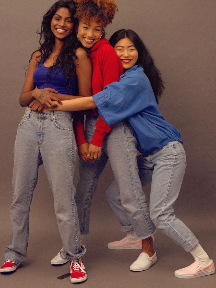 three young women are posing for a photo.  the woman on the left is wearing jeans, a blue tank top, and red sneakers.  the woman in the middle is wearing jeans, a red sweater, and white sneakers.  the woman on the right is wearing jeans, a blue shirt, and white sneakers. they are all smiling and appear to be close friends.