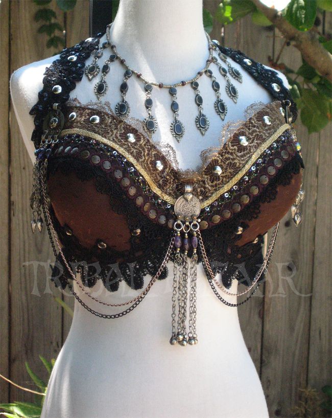 a close up view of a ornate belly dancing bra on a white mannequin. the bra is made of brown, black, and gold fabric and features a variety of beads, chains, and other embellishments. the bra is very intricate and detailed, with a lot of different textures and patterns.  the bra is on a white mannequin and the background is blurred.