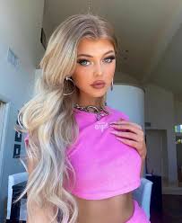 a young woman, loren gray, with blonde hair, is posing for a selfie indoors. she is wearing a pink crop top, hoop earrings, and a necklace. she has blue eyes and is wearing makeup.