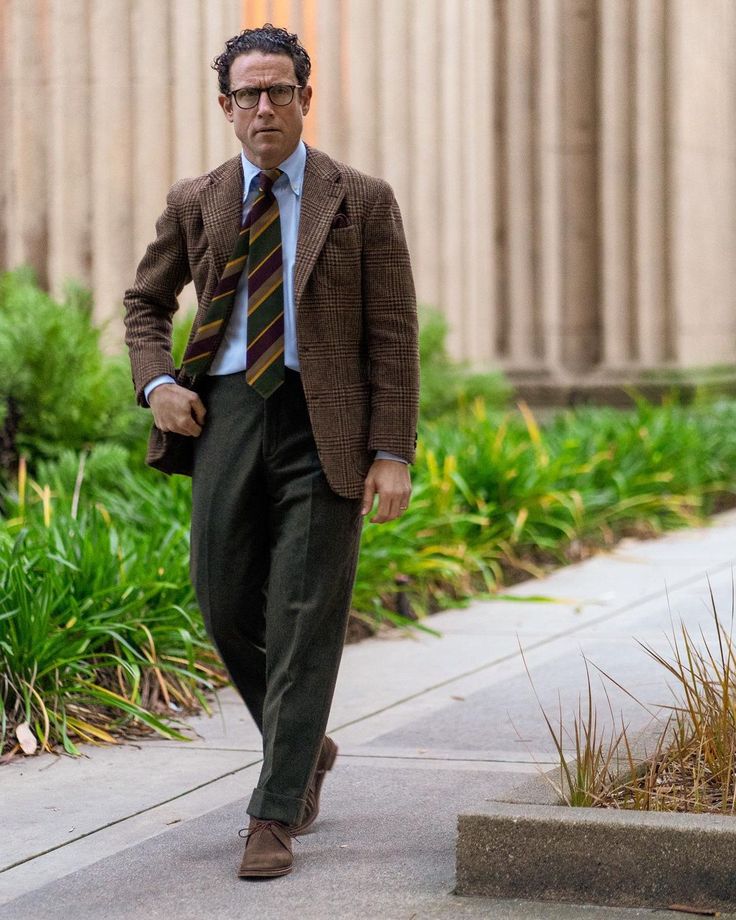 a man in a brown tweed jacket, green trousers, and brown suede shoes walks down a pathway. he's wearing a light blue shirt and a striped tie, and eyeglasses. the setting is outdoors, with landscaping featuring plants on either side of the path. the overall style is classic and formal.
