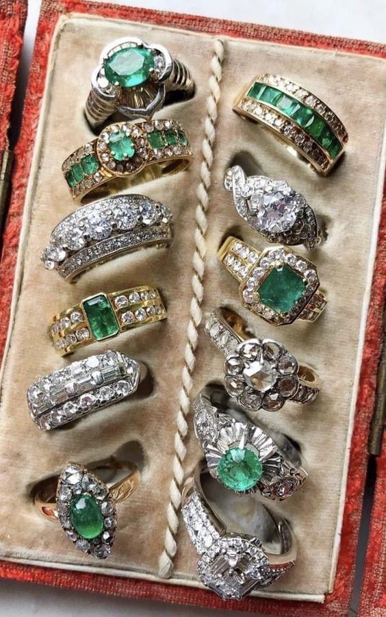 a closeup shot of a collection of rings with emerald gemstones. the rings are arranged in a red velvet box, showcasing their sparkle and elegance. the focus is on the emerald gemstones, which are surrounded by diamonds and other embellishments. the image is a testament to the artistry and craftsmanship involved in creating these exquisite pieces of jewelry.