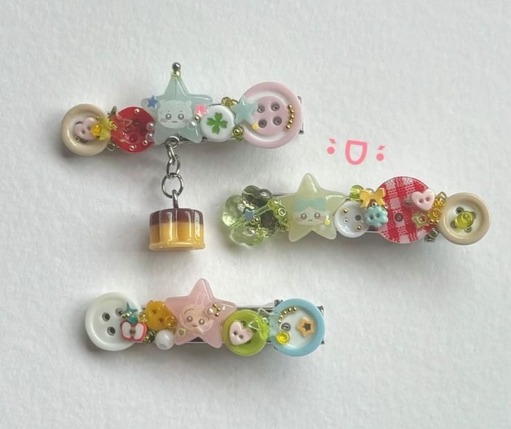 three kawaii hair clips are displayed on a white surface. each clip is uniquely decorated with an assortment of buttons, starshaped charms, beads, and other small embellishments. the topmost hair clip features a hanging charm resembling a small pudding.
