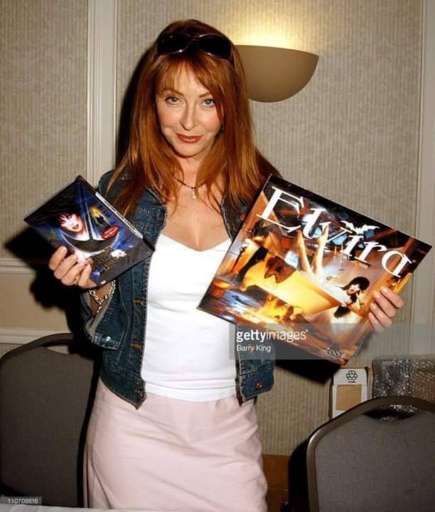 a woman with long dark hair, wearing a denim jacket over a white shirt and a pink skirt, is smiling and holding a book in her hands. she is signing the book for a fan at a convention. she is standing indoors, looking at the camera.
