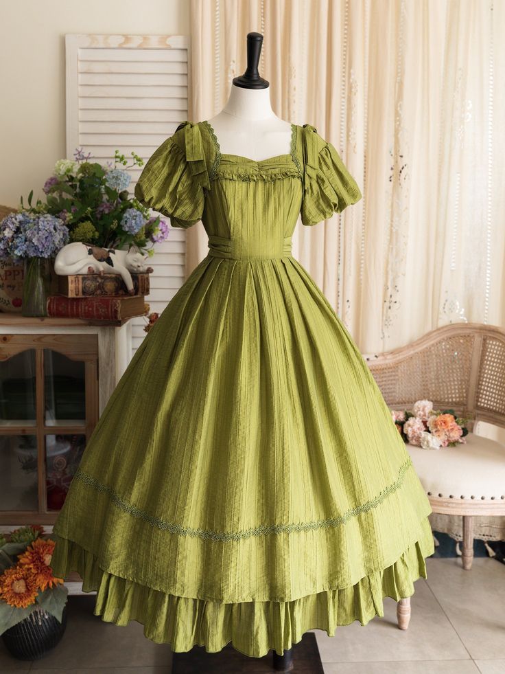 a green vintageinspired dress is displayed on a mannequin. the dress features puff sleeves, a square neckline, and a flowing tiered skirt with lace trim. the background includes a white wall, a floral curtain, a wooden cabinet, and flower arrangements.