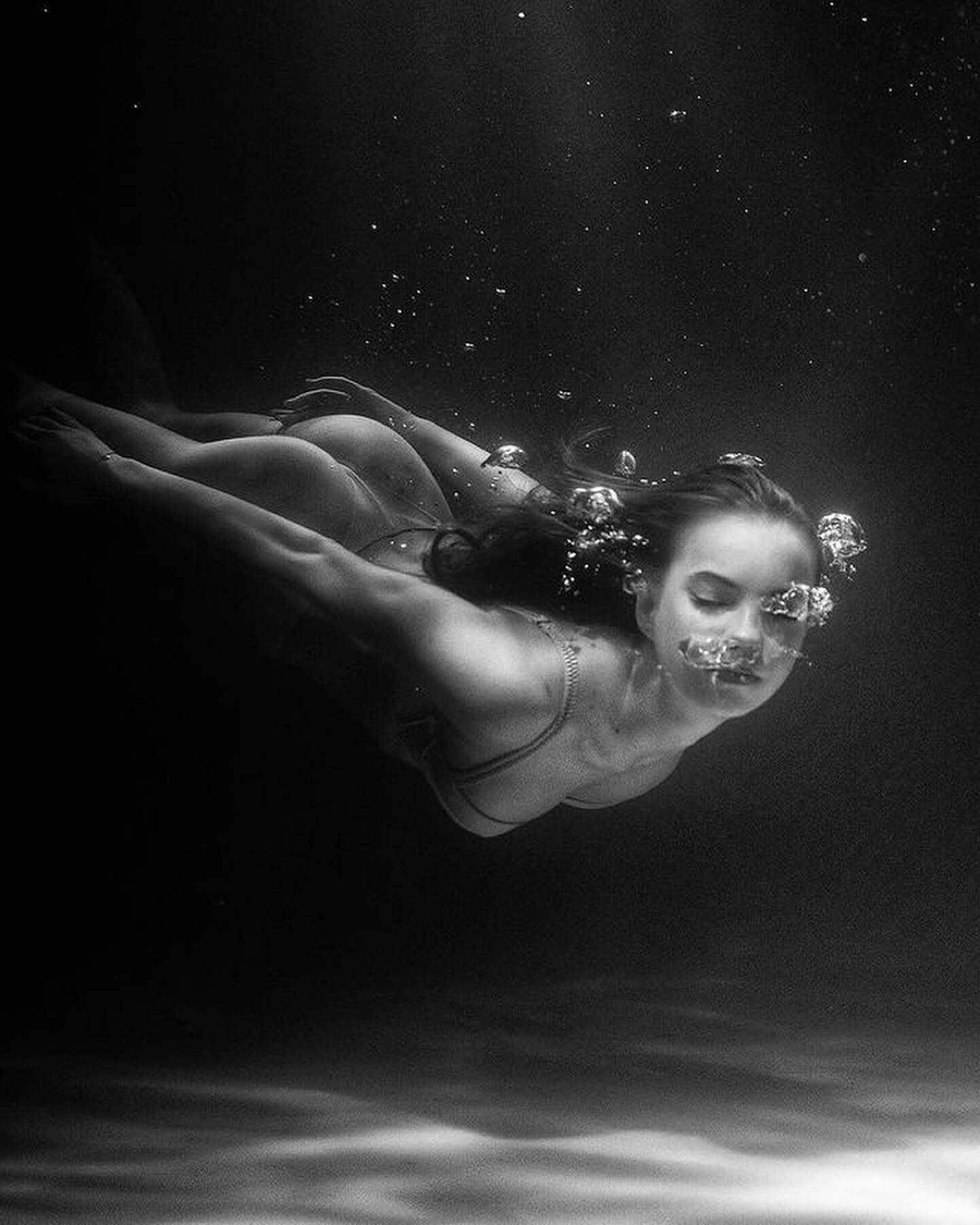 the image is a black and white underwater photograph of a woman swimming.  she's submerged in dark water, her body angled slightly upwards.  air bubbles surround her face and rise towards the surface. the woman is wearing a darkcolored bikini top, and her long hair flows behind her. the overall mood is serene and elegant, with the contrast between the dark water and the lighter tones of the woman's body creating a dramatic effect. the lighting highlights her form and features, drawing the viewer's eye to her peaceful expression. the background is dark and indistinct, keeping the focus on the woman.