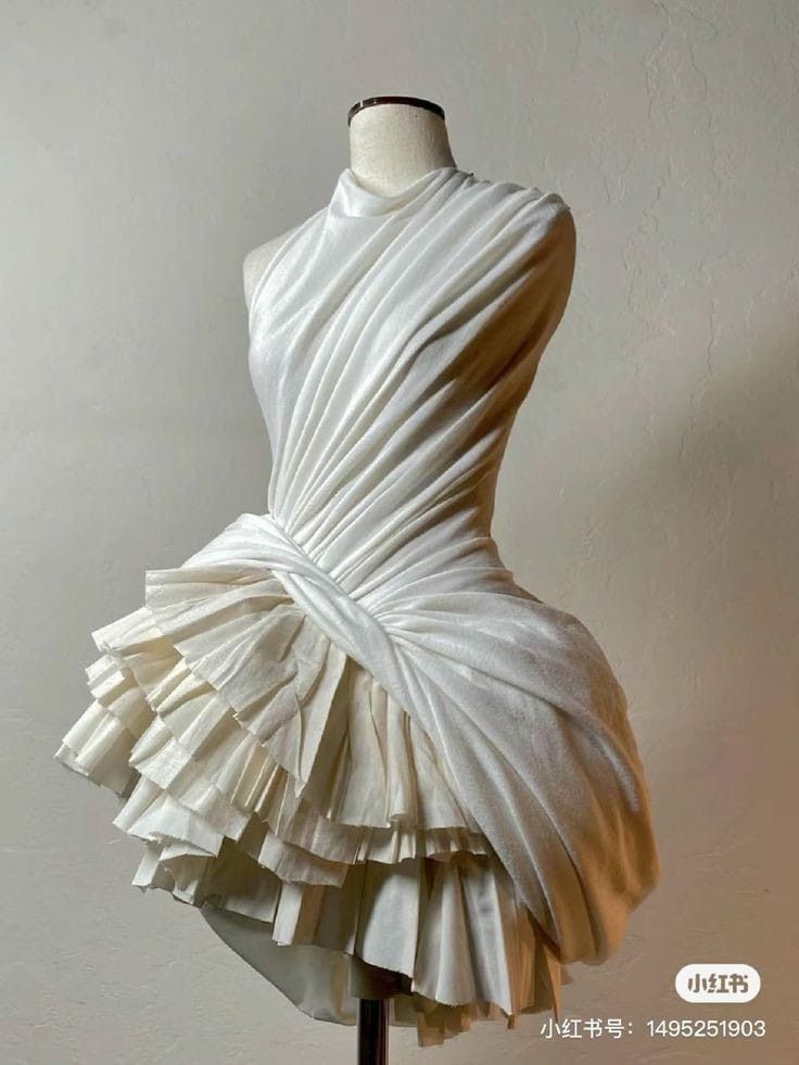 a white dress is displayed on a mannequin. the dress features a high neckline, draped fabric across the bodice, and a layered, pleated skirt with an asymmetrical hem. it is a fashion design piece, potentially couture.