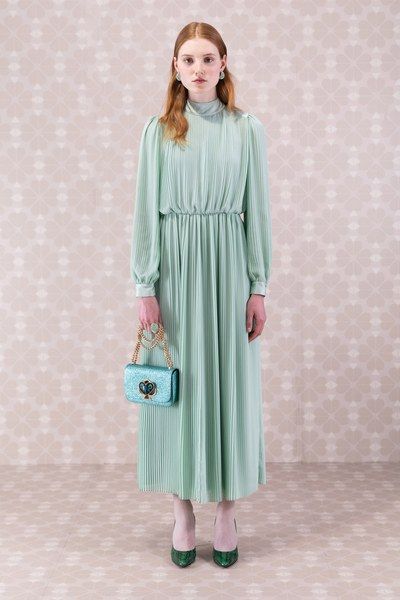 a fulllength studio shot of a woman wearing a light green pleated maxi dress. she is holding a light blue kate spade handbag and wearing green high heels. the background is a light floral pattern. the overall style is elegant and feminine.