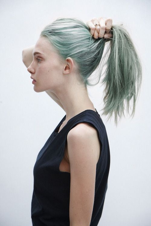 a woman with green hair is shown in profile, facing left, holding her hair in a ponytail. she is wearing a black tank top.