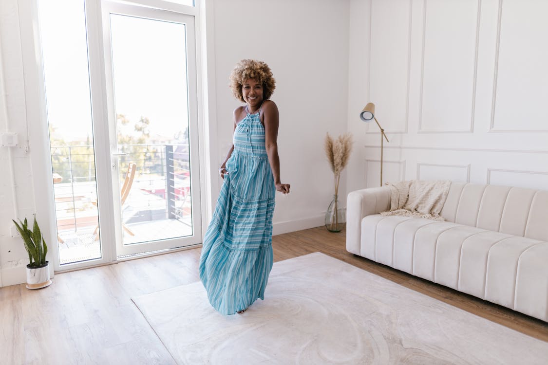 a young black woman with curly blonde hair is joyfully dancing in a light blue and white striped maxi dress in a bright, modern living room.  she's smiling happily. the room is decorated in a minimalist style with a white sofa, white walls, and large windows letting in abundant natural light. a small plant sits on the floor next to the window, and a floor lamp stands near the sofa.  the overall mood is cheerful and carefree.