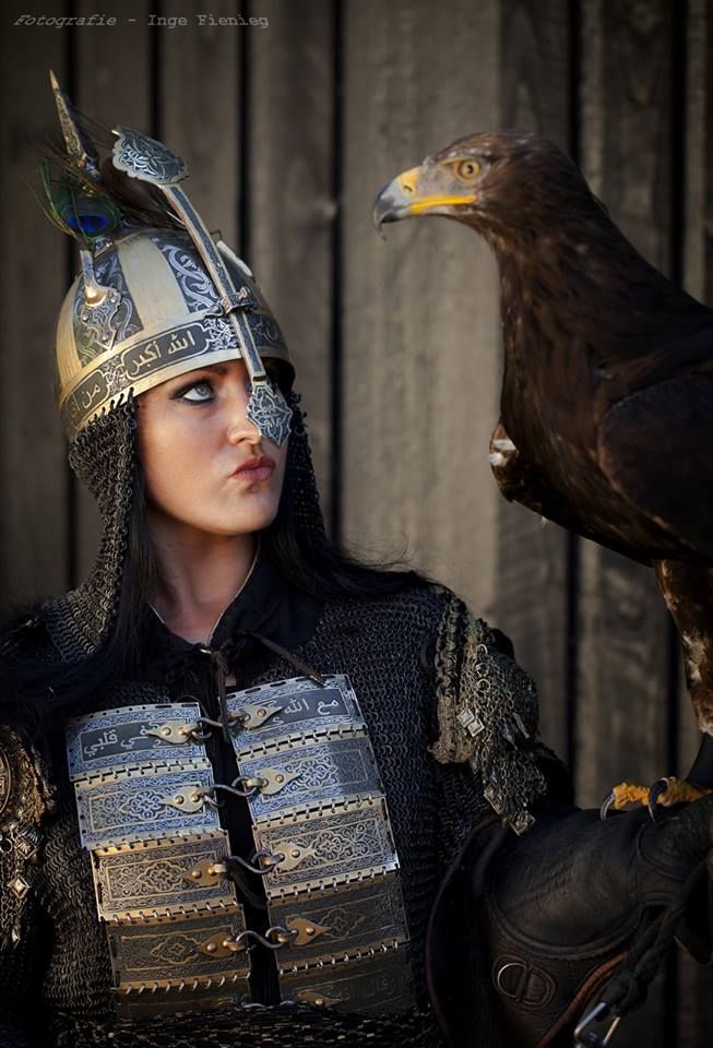 a woman in armor with a helmet and chainmail stands next to an eagle. the woman is looking at the eagle with a slight pout on her lips. the eagle is perched on her gloved hand. the helmet has arabic text on it. the armor is decorated with metal plates and chainmail.