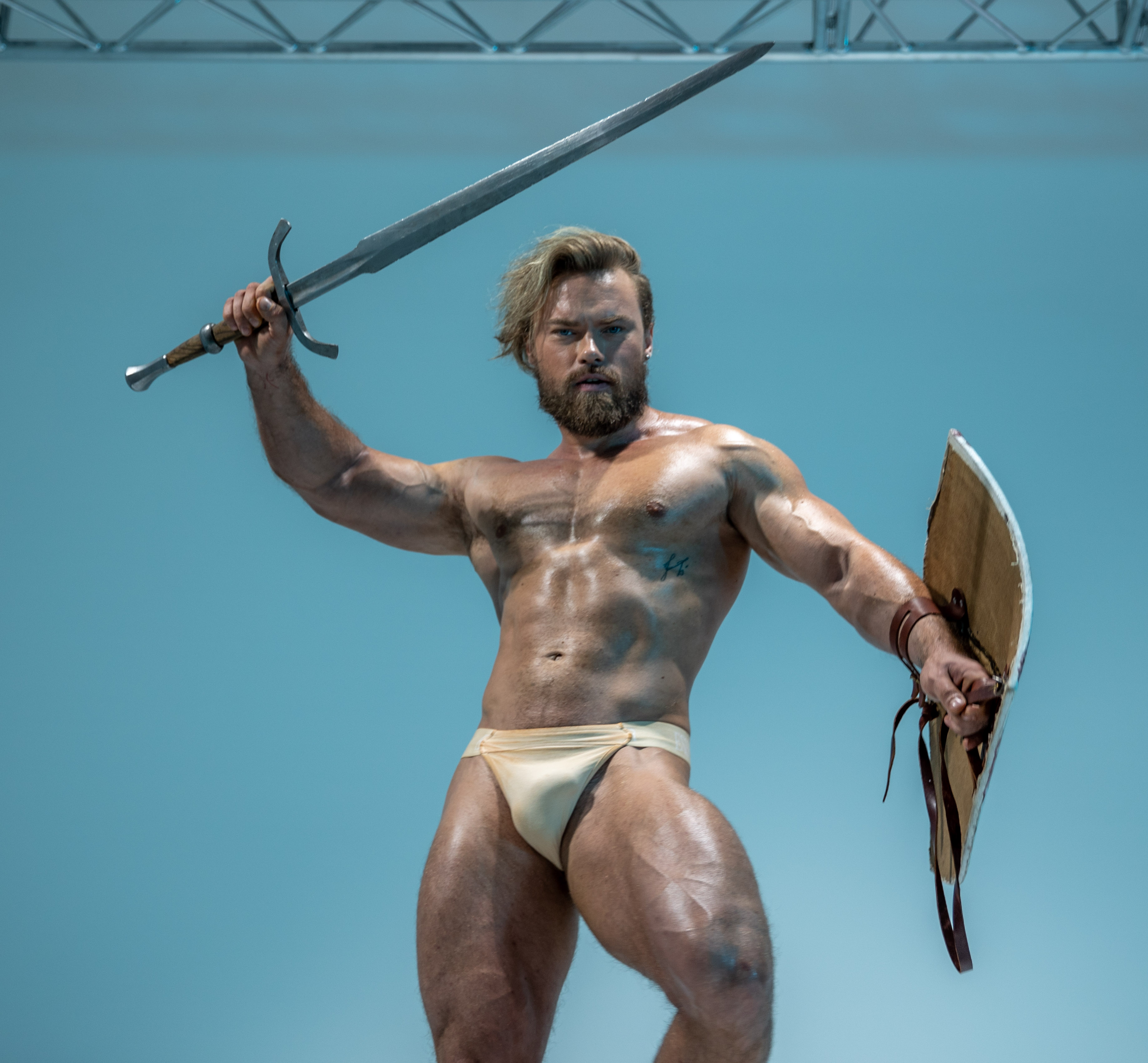 a muscular man with a beard and long blond hair, styled to appear like an ancient warrior,  stands against a light blue background in a fighting stance. he holds a sword in his right hand, raised above his head, and a shield in his left.  he is wearing a beige loincloth. the image is shot from a low angle.