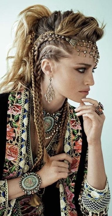 a woman with braids and various pieces of jewelry is shown in a side view. she is wearing a floral patterned jacket and has her hand near her face.