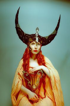 a woman with long red hair wearing a flowing peachcolored dress sits holding a golden goblet. she is adorned with an elaborate headdress featuring large black horns embellished with silver details. the image has a mythical, goddesslike quality. the background is a pale teal, and the lighting is soft and ethereal.