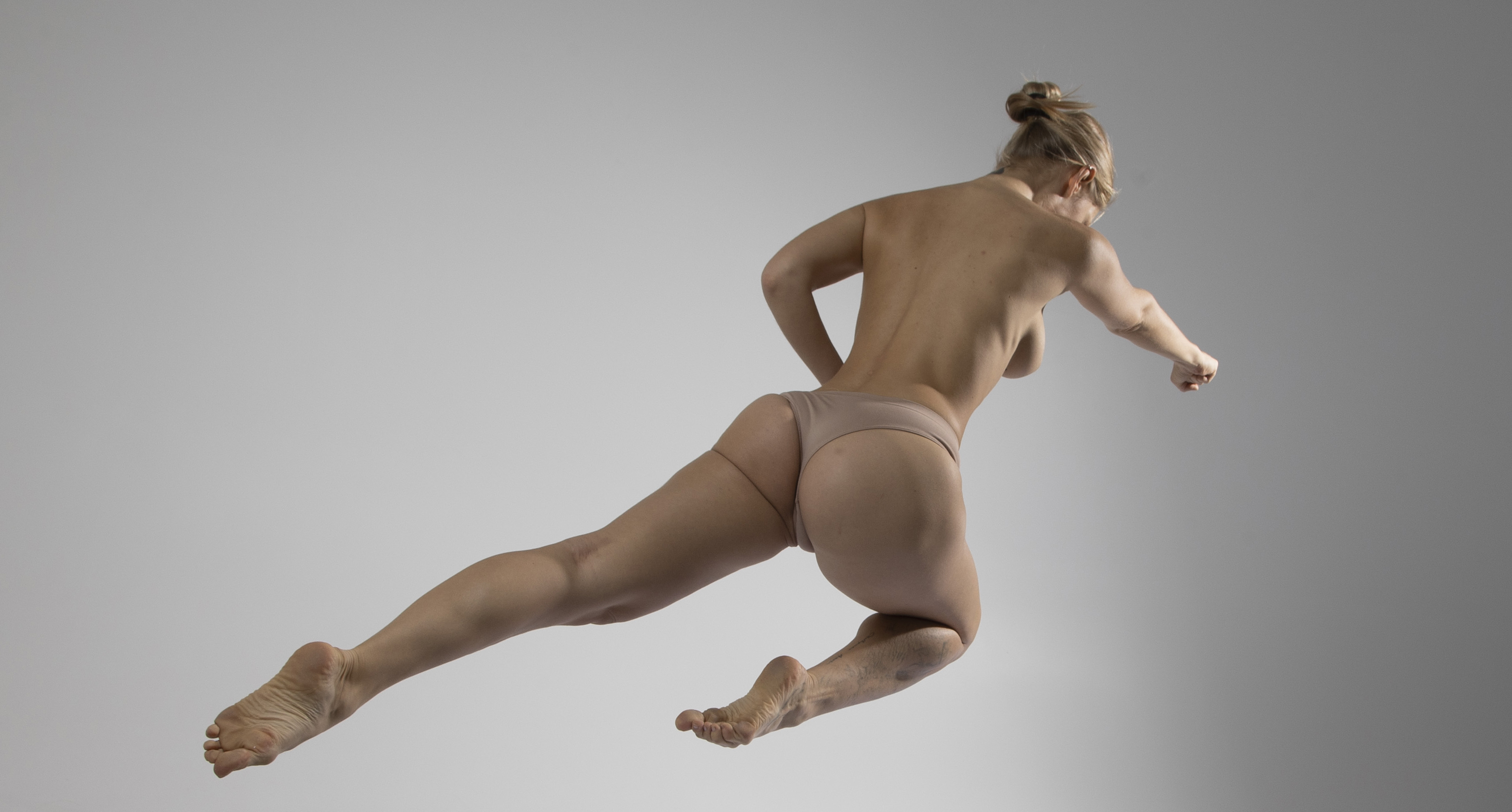 a nude woman is captured in a dynamic studio shot against a gray background. she's in midair, executing a punch with her right arm extended, while the rest of her body reflects the motion of a jump. the photograph focuses on her back and highlights her athletic physique.