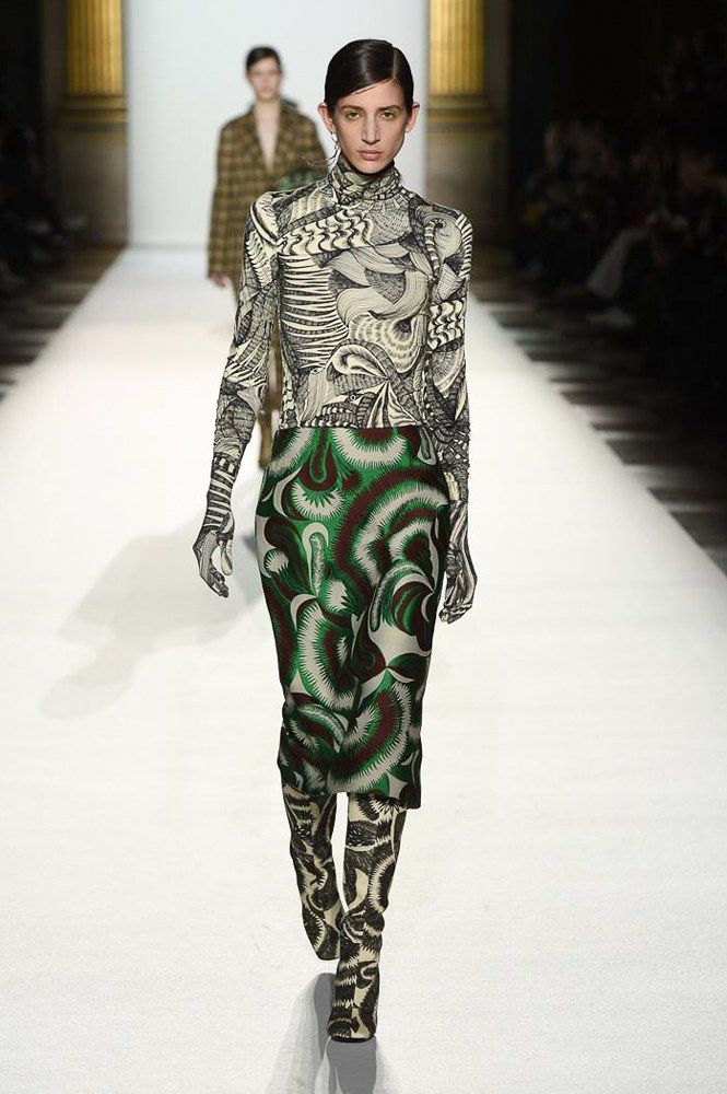 the image shows a female model walking down a runway during a fashion show. she is wearing a fitted, kneelength skirt and a matching longsleeved turtleneck top, both featuring a striking abstract print in shades of green, black, and white.  the pattern is bold and swirling. she's also wearing high, patterned boots that complement the outfit's print. the background is blurred but reveals a typical runway setting with other models and onlookers. the overall impression is one of high fashion and contemporary style.