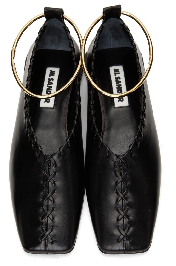 a closeup overhead shot of a pair of black leather jil sander flats.  the shoes have a square toe, a crisscross stitching detail at the front, and are adorned with a gold hoop that acts as an ankle strap.  the jil sander logo is visible inside each shoe.  the shoes are presented against a plain white background.