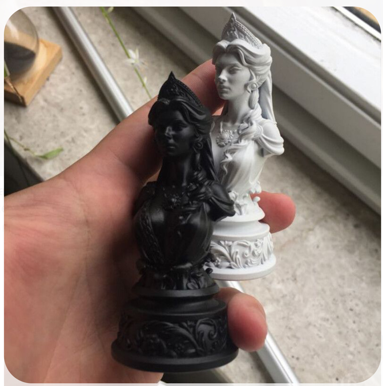 a person is holding two 3dprinted busts of a queen, one in black and one in white, both with intricate details. the busts resemble chess pieces. the background is a window with a blurry view of the outdoors.