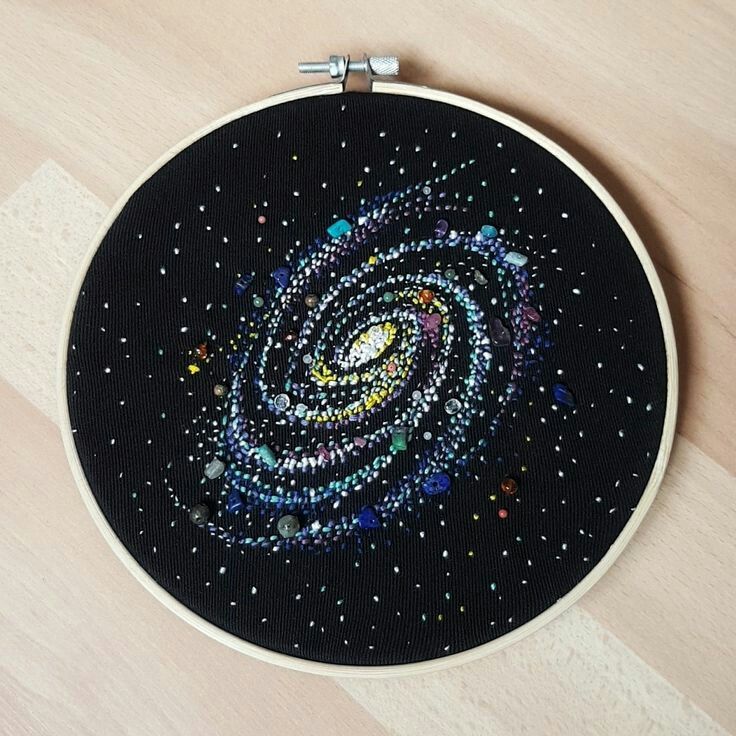 an embroidery hoop displays a handmade galaxy design on black fabric. the design features a spiral galaxy with embroidered stars and the use of colorful beads.