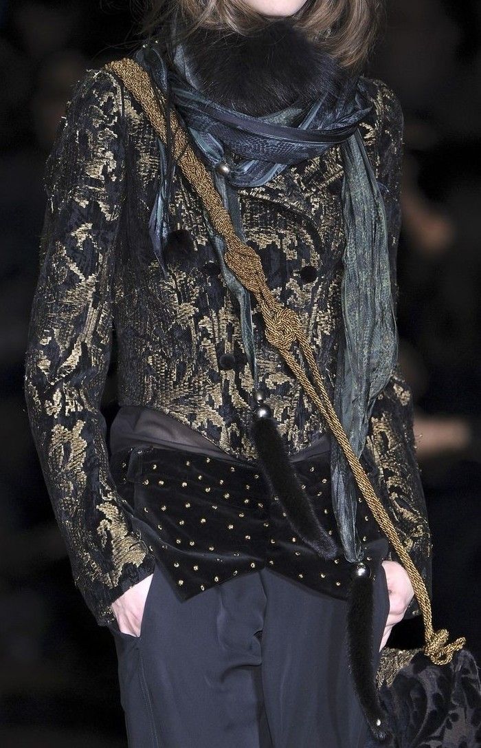 a female fashion model wears a black and gold patterned jacket with a fur trim and a long scarf. she is standing with her hands in her pockets and looking to the side.