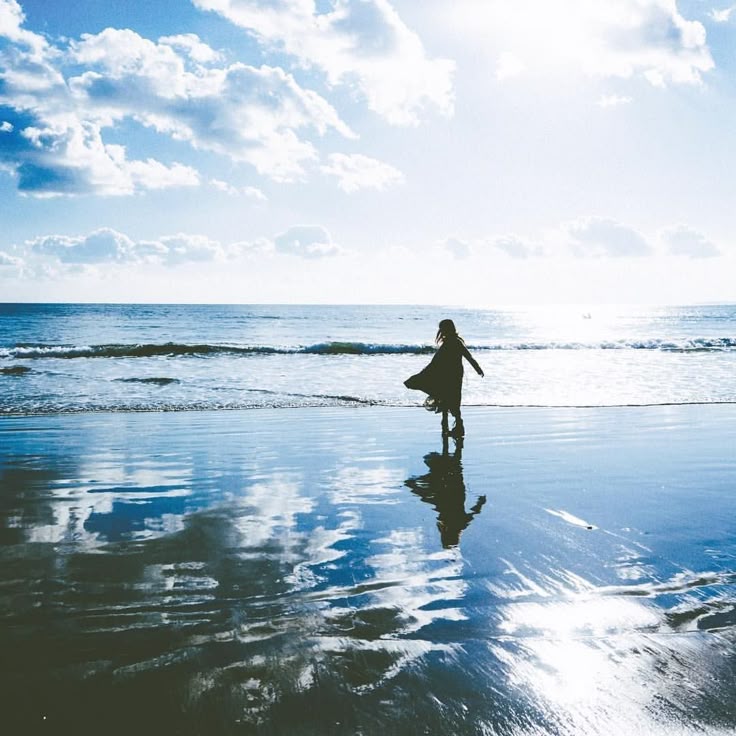 a woman stands on a beach with shallow water reflecting the sky and clouds. the sky is bright blue with fluffy white clouds. the ocean stretches out to the horizon, where the sunlight creates a bright glare. the woman is silhouetted, wearing a dress that flows in the breeze. her reflection is visible in the wet sand.