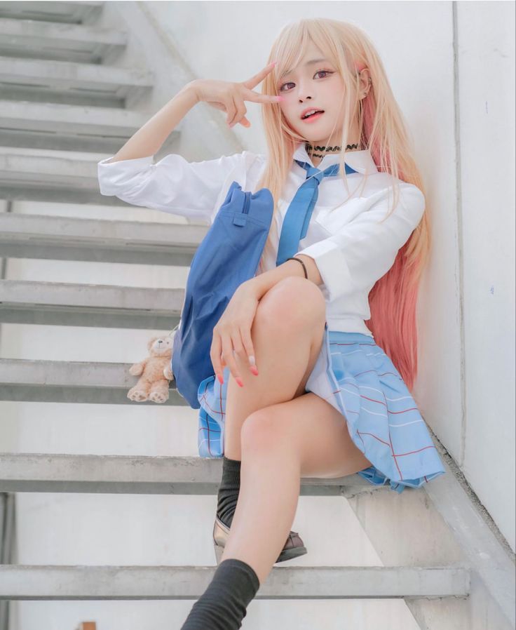 a young woman in a schoolgirl cosplay outfit sits on a set of stairs. she has long pink hair, and she is wearing a white blouse, a blue plaid skirt, black socks, and black shoes. she is making a peace sign with her hand, and she looks playful and happy. the background is a light gray wall and the stairs are metal. a small stuffed animal sits next to her on the stairs.