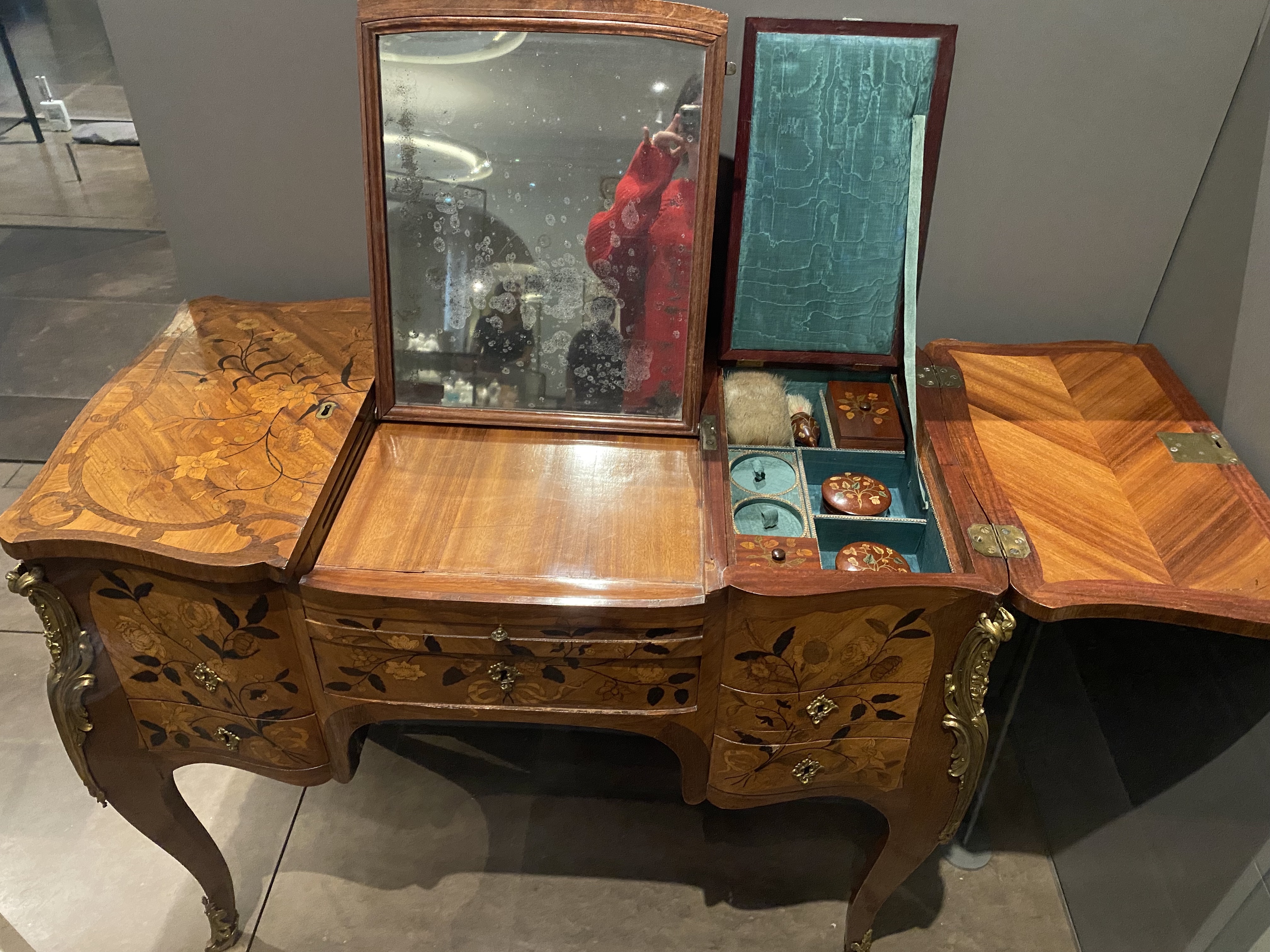 a detailed image shows an antique dressing table with ornate wood inlay and marquetry. the table features a central section with a mirror and multiple drawers, flanked by two hinged sections that open to reveal storage compartments. the interior of the right compartment is lined with green velvet, and contains various decorative objects and possibly cosmetics. the table is richly decorated with floral motifs and brass fittings, supported by ornate curved legs. the mirror reflects a person in red clothing.