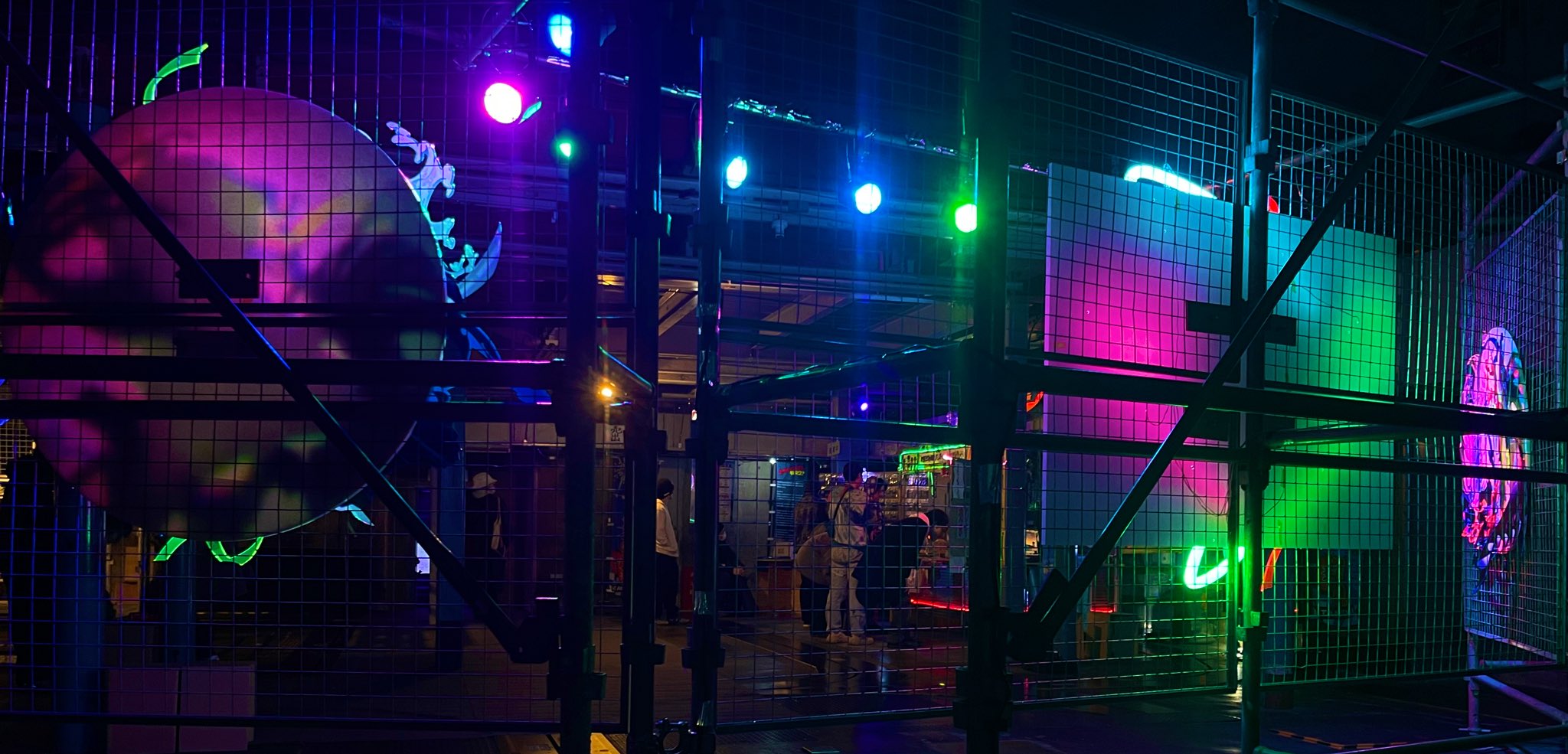 this vibrant image captures the electric atmosphere of a live music event. taken from behind a metal fence, likely a barrier separating the audience from the stage, it offers a glimpse into a club or concert venue. colorful stage lights in shades of purple, green, and white illuminate the space, creating an energetic and immersive experience. silhouettes of audience members and partially obscured performers suggest a lively and engaging performance. the overall impression is one of excitement, energy, and the thrill of live entertainment.
