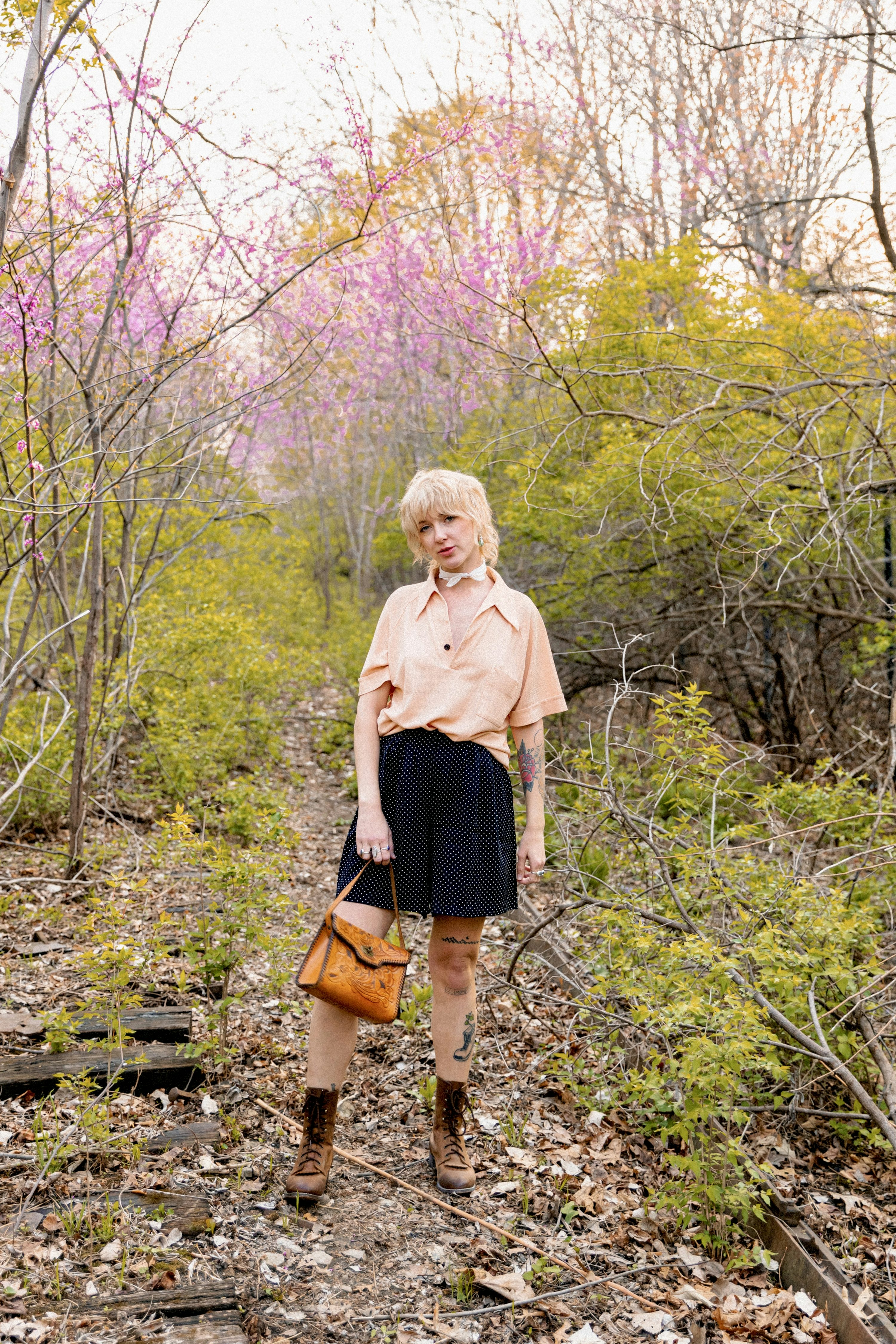 a stylish blonde woman in a peach vintage blouse, black polka dot shorts, and brown boots poses on a path in a forest with pink and yellowgreen flowering trees. she carries a brown leather handbag.