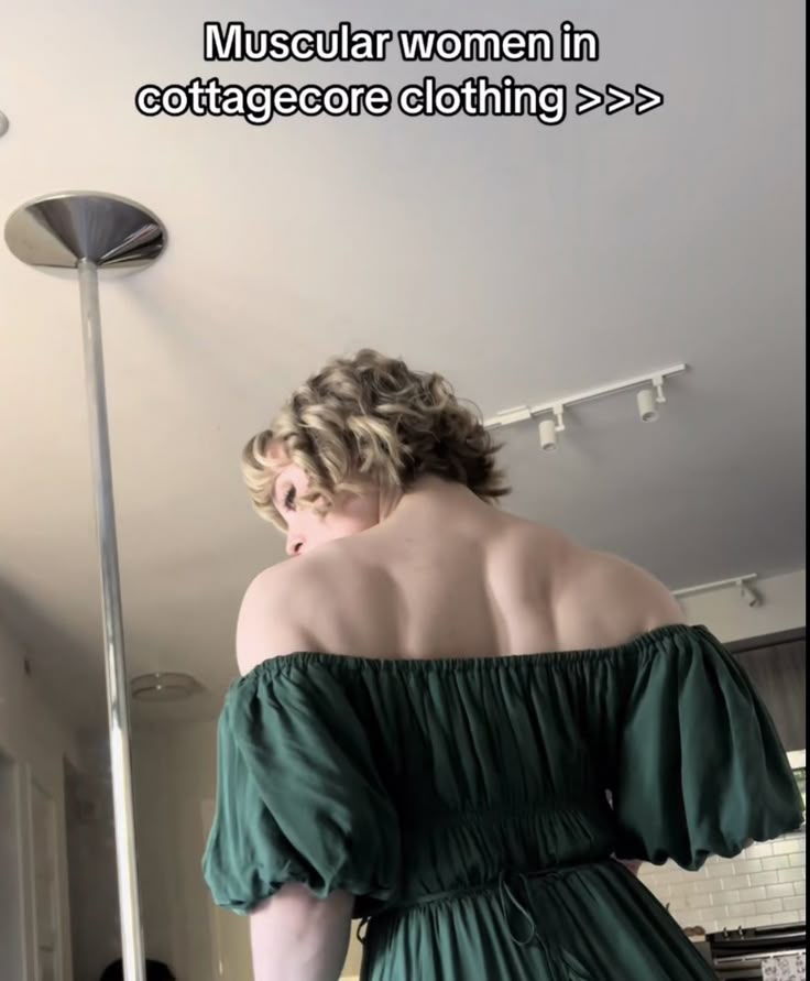 a woman with a muscular back and blonde hair wears a green offtheshoulder cottagecore dress. the text overlay reads "muscular women in cottagecore clothing >>>". the scene is indoors with a ceiling lamp visible.