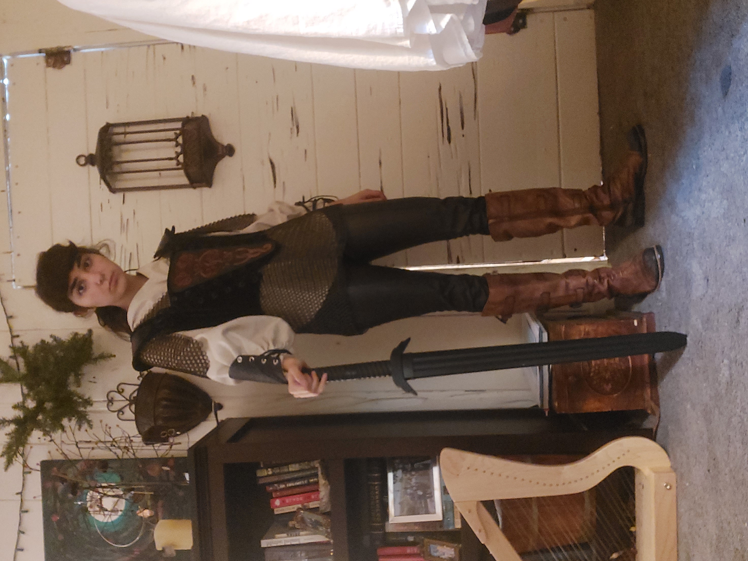 young woman dressed in a medieval fantasy costume stands holding a large sword. she is wearing a chainmail vest over a white shirt, dark pants, and brown boots. her hair is pulled back. she is posing in a room with a wooden door and a bookshelf behind her. a harp is visible to the left, and a wooden box is at her feet. the overall scene evokes a fantasy setting.