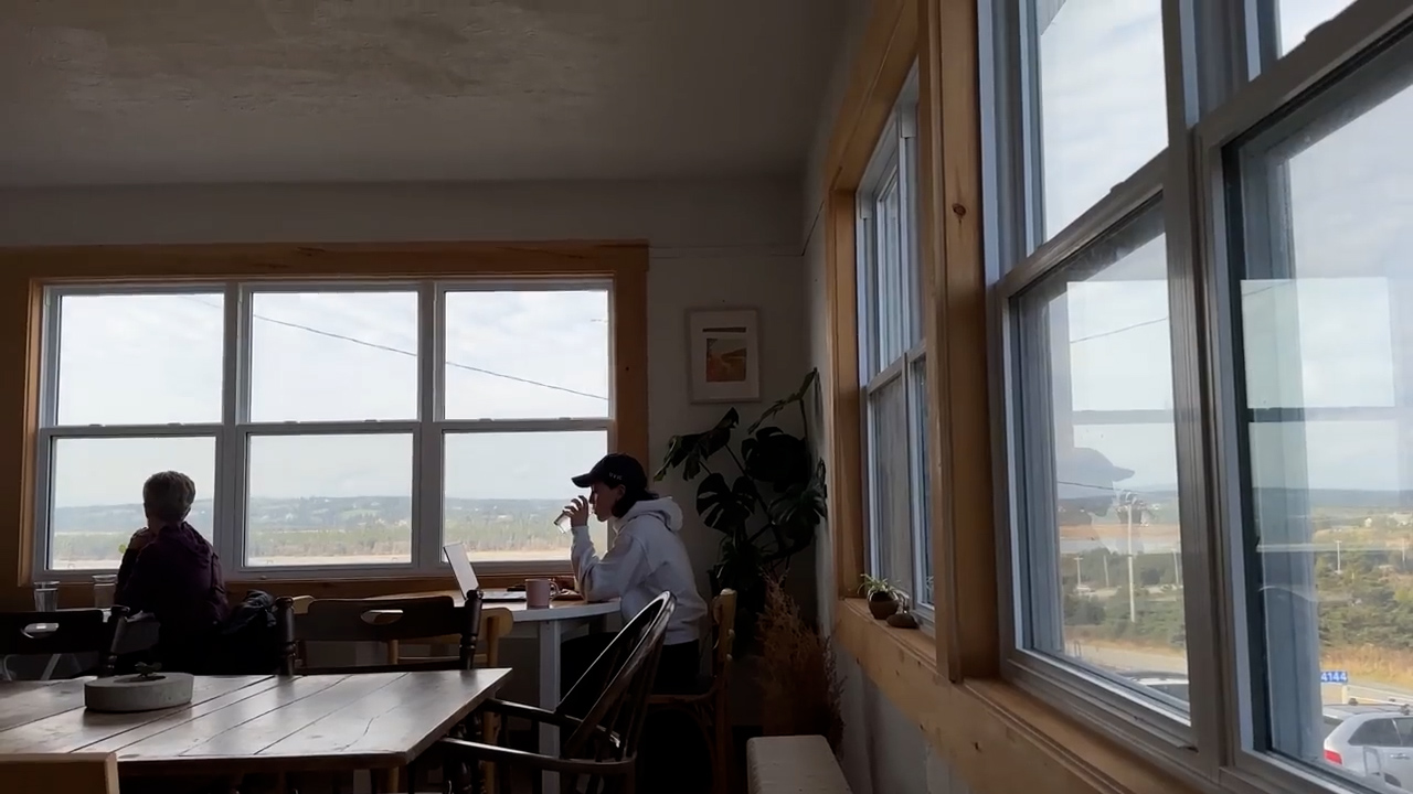 a photo of the interior of a cafe with large windows that look out onto an overcast sky and a distant town. one woman sits with her back to the camera, seemingly looking out the window, and another sits facing the window at a small white table working on a laptop. the cafe is decorated with light wood and a few plants. the overall feeling is bright and airy, but calm and quiet.