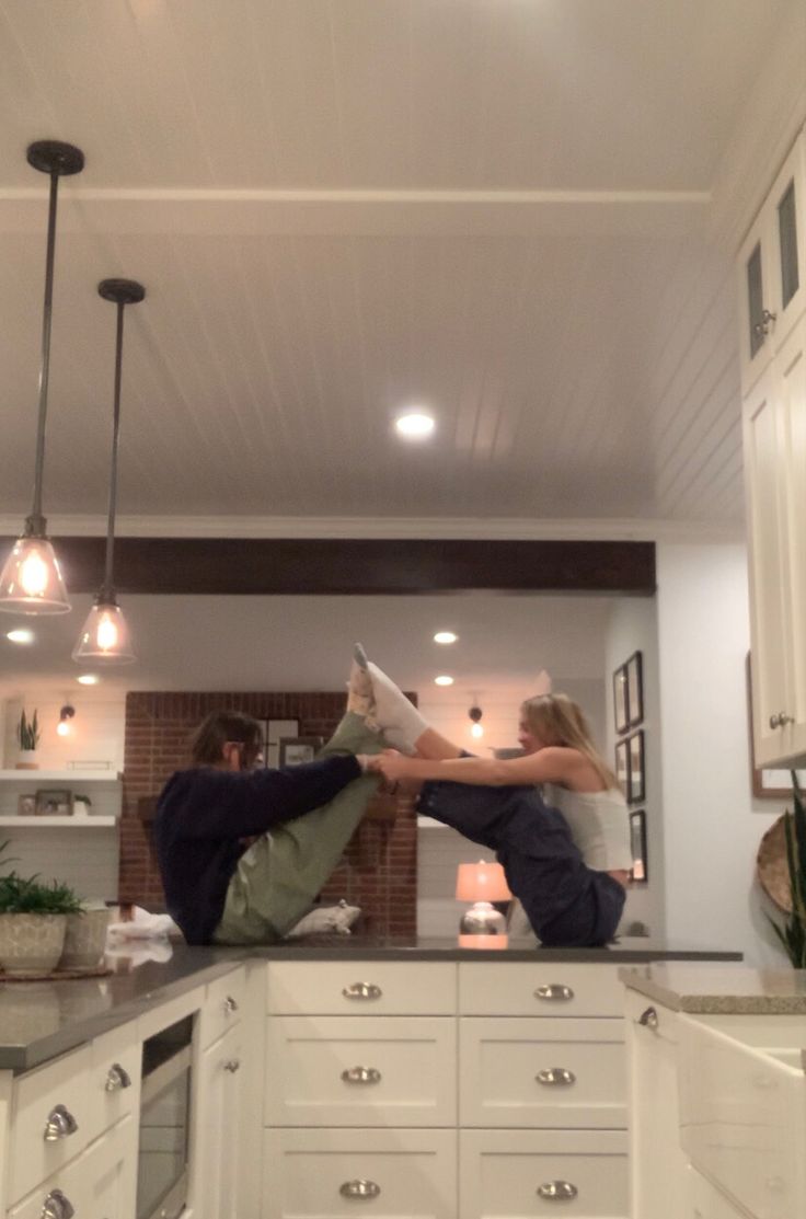 a couple is playing around in the kitchen. they are both standing on the counter, and the girl is holding the boy's legs up in the air. they are both smiling and seem to be enjoying themselves. there are white cabinets and drawers below the counter, and a sink and faucet to the right. the room is decorated in a modern and minimalist style.