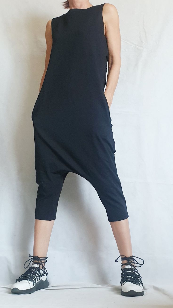 a woman is wearing a black jumpsuit and white sneakers with black laces. she is standing and posing for the camera.
