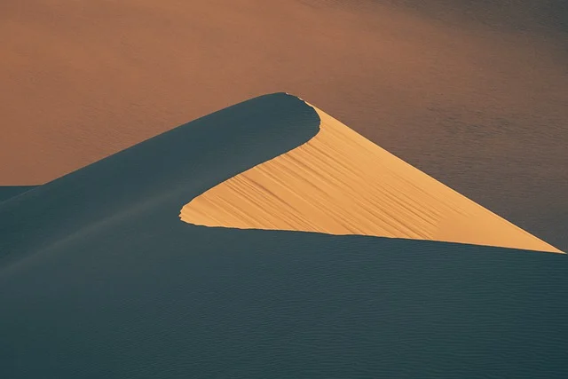 the image showcases a minimalist view of a sand dune in the desert, captured from an aerial perspective. the lighting suggests it's near sunset, casting long, cool shadows on the windswept sand. the palette features warm hues of sand contrasted against the cool blues of the shadowed areas.  the dune's sharp, cresting form creates an almost abstract and graphic element against the vastness of the desert landscape.