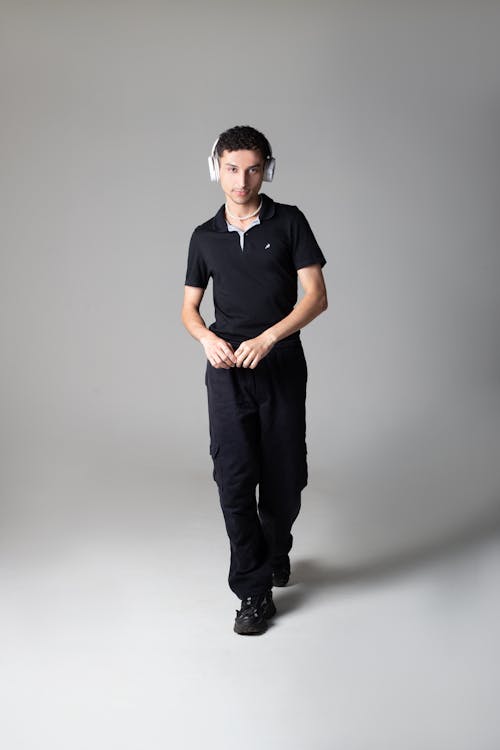 a young man wearing black casual clothing, including a polo shirt and cargo pants, is walking towards the camera. he has on white overtheear headphones and is looking directly at the camera.  the background is a plain light grey. the overall style is simple and uncluttered.