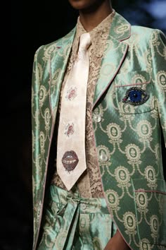 a closeup photo of a woman wearing a stylish, green patterned jacket with a floral patterned tie with a red lip design on it. the woman's outfit is elegant and stylish, with a lace top peeking out from beneath the jacket. the jacket has an ornate design and looks highquality and expensive.