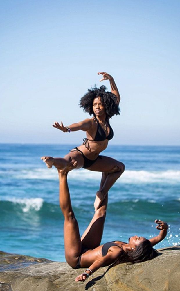 two women pose in an acrobatic yoga pose on a rock on a beach. they are both wearing bikinis and have tan skin. the woman on the bottom is supporting the woman on top with her feet. the woman on top is in a dynamic pose with her arms extended. the ocean and waves are in the background.