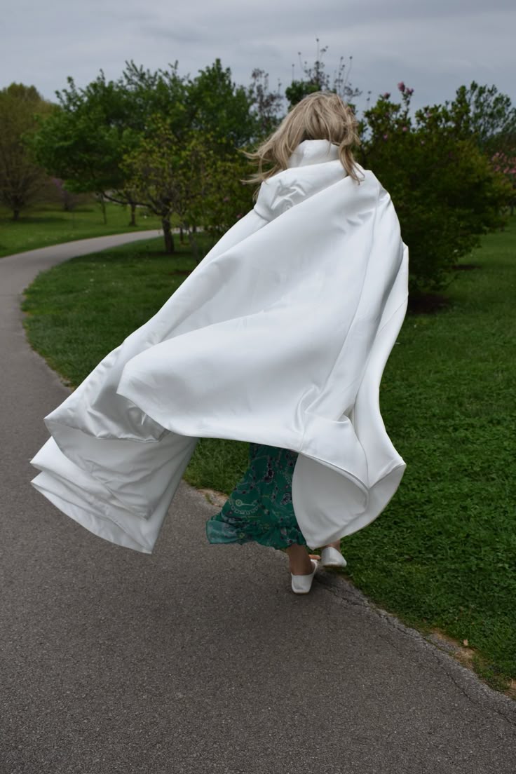 a woman with blonde hair is seen from behind, walking on a gray asphalt path with green grass on either side. she's wearing a large, flowing white cape that is caught by the wind, billows around her. beneath the cape, a green dress with a busy pattern is visible. her feet are clad in white shoes. the background features several green trees under an overcast sky, suggesting a park setting. the scene conveys movement and a sense of freedom.