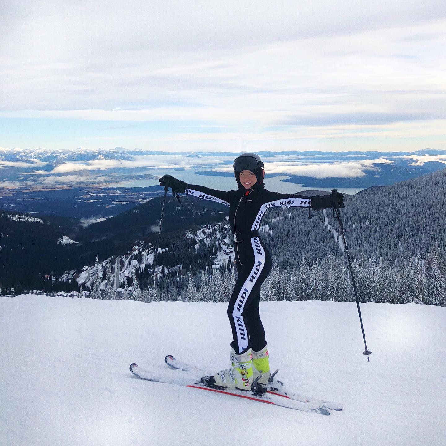 a woman in a black ski suit with white lettering is standing on a snowy mountaintop with her skis and poles. she is smiling and looking at the camera. the background shows a stunning view of snowcovered mountains and a vast expanse of land. the sky is partly cloudy, and the overall image conveys a sense of winter sports adventure and beauty.