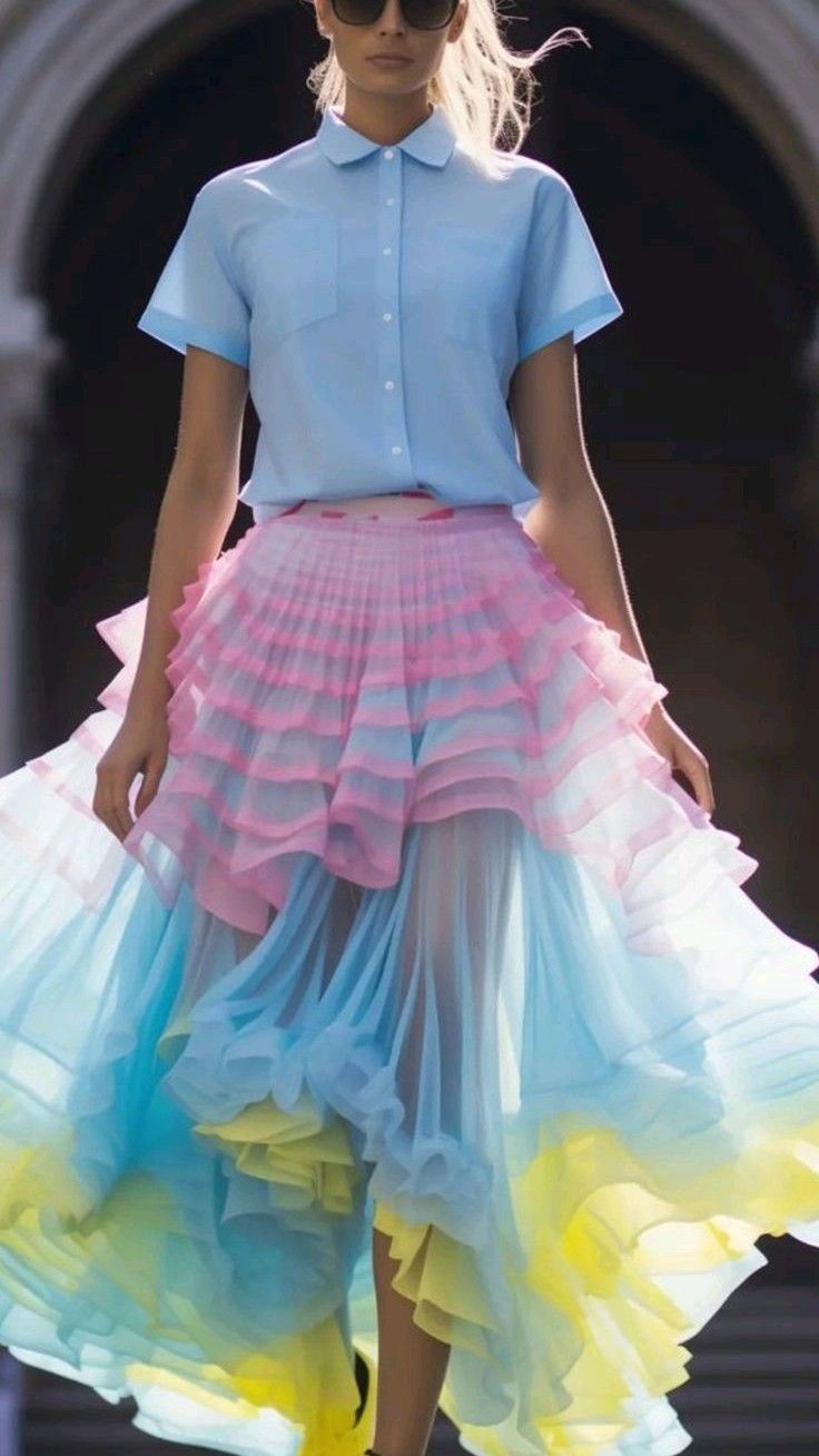 a woman in a stylish outfit is standing in a street, her back to the camera. she is wearing a pale blue buttondown shirt, a layered pink and blue tulle skirt with yellow accents, and sunglasses.
