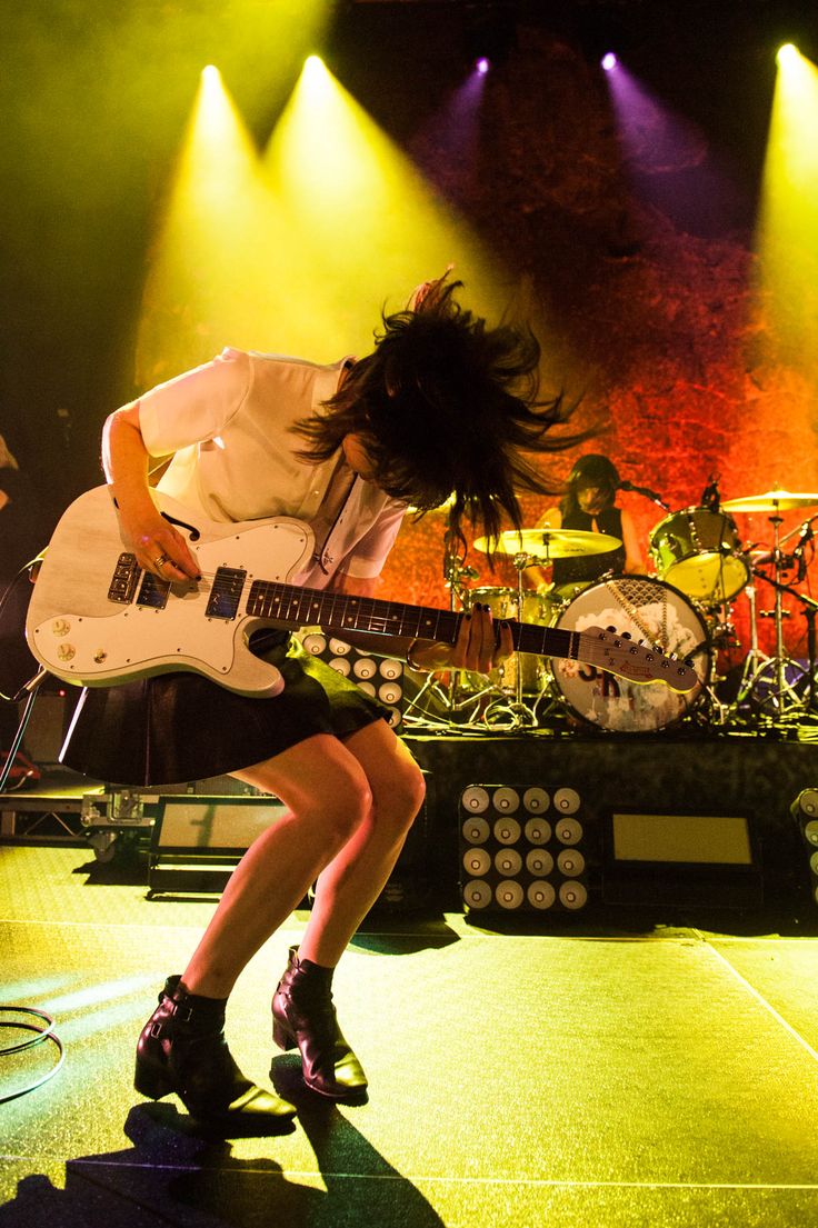 a female guitarist in a white shirt and black skirt is shown midperformance on a stage with spotlights, performing for a crowd. she's playing a white electric guitar and the crowd is clearly enjoying her performance.