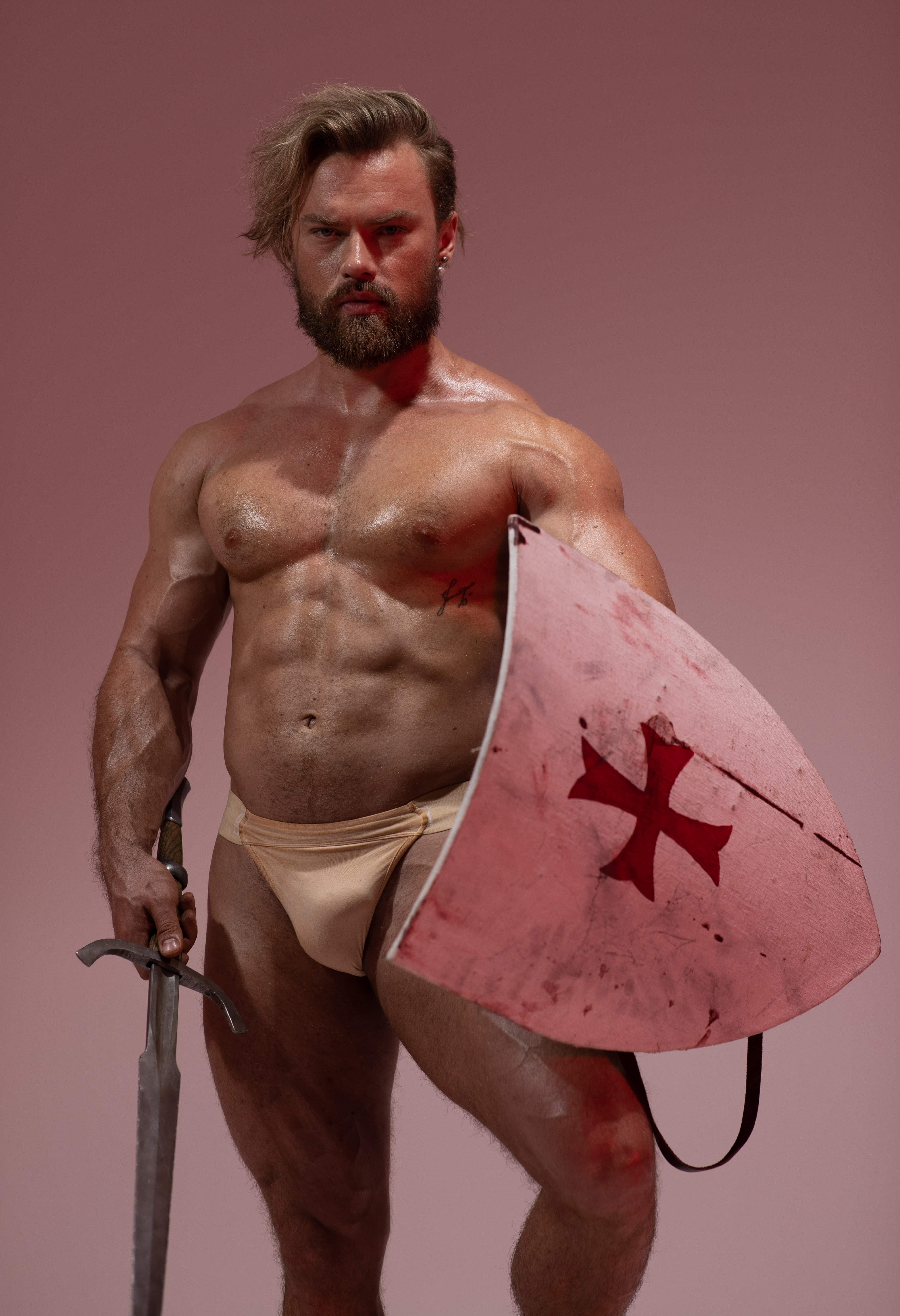 a muscular man with a beard and long hair pulled back stands against a pale pink backdrop. he is dressed as a warrior, wearing only a loincloth and holding a sword in his right hand and a large, pink shield in his left. the shield features a red cross. he has a powerful and imposing presence. the lighting is dramatic, highlighting his musculature and creating a sense of strength and power.