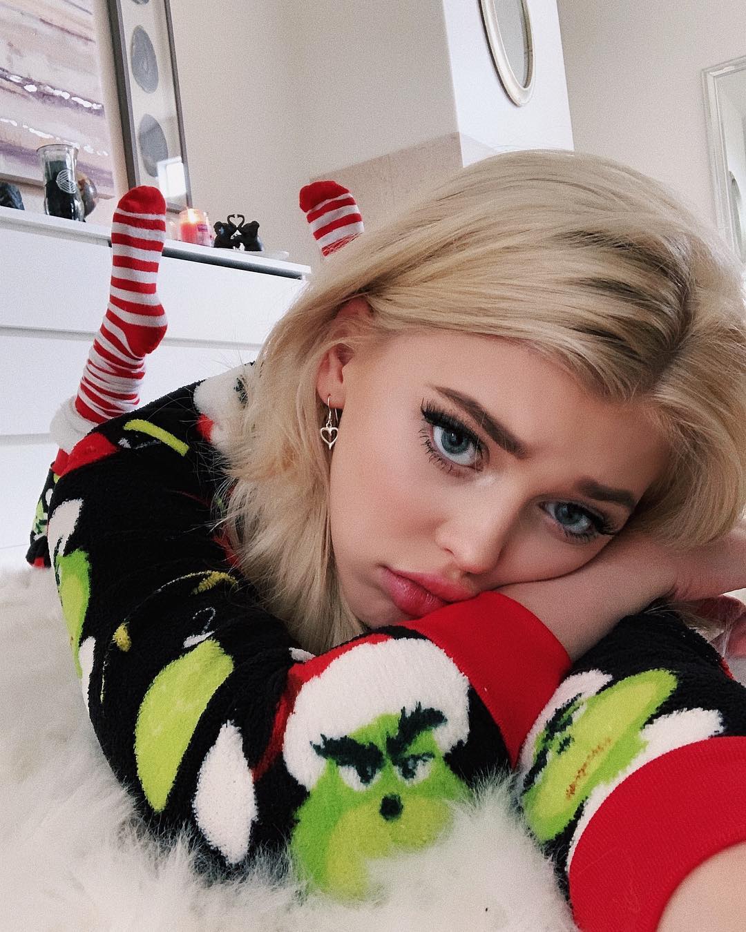 a woman with blonde hair and blue eyes is lying down on a white rug. she is wearing a black sweater with grinch designs on it and red and white striped socks. she has heart earrings. behind her is a dresser with a picture frame and candle on top.