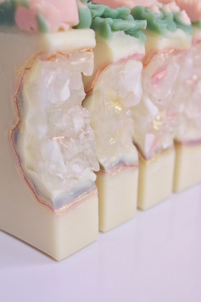 a closeup shot of four bars of soap in a row. the soap is white with a pink and green swirl on top. the soap is also decorated with gold glitter and crystals.  the soap is arranged on a white surface. the soap is likely handmade and natural, with a delicate floral fragrance.