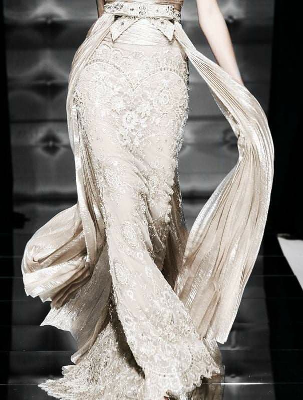 a model walks the runway in a beautiful white dress, the dress is adorned with lace, and has a long train. the dress is very elegant and glamorous, and it is clear that this is a highfashion garment. the model is wearing her hair up, and she has a natural makeup look.