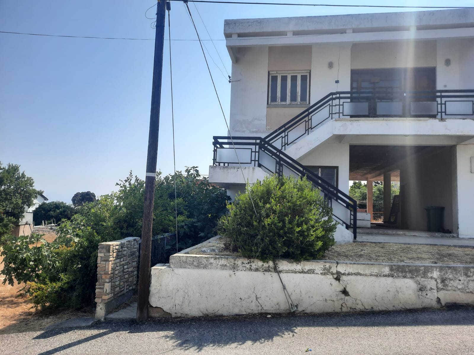 the image shows a twostory house with an exterior staircase, a white facade, and black metal railings, situated beside a street with utility pole on the edge. there are shrubs and a wall in the foreground, and the sky is visible in the background.